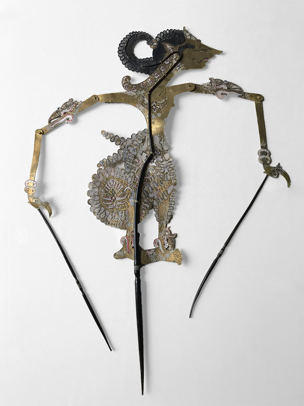 Marioneta de cuero calado y pintado que representa a un personaje de perfil. Pueden verse la varilla central que sostiene su cuerpo y las varillas en cada mano para realizar los movimientos de los brazos articulados.